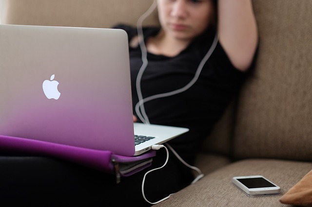 Dievča so slúchadlami v ušiach leží na gauči a pozerá do notebooku